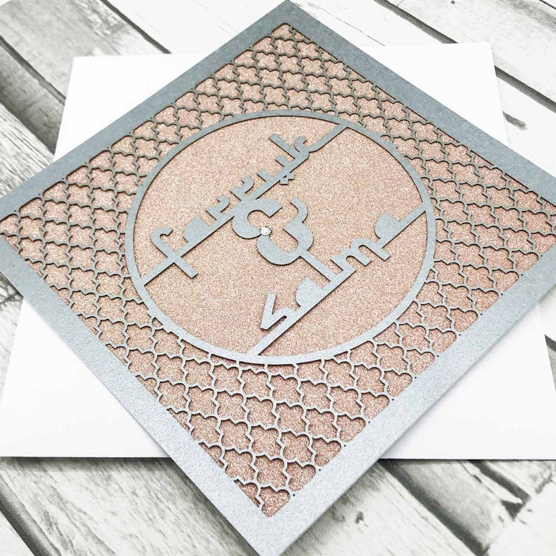 K&ouml;nnte beinhalten: Eine lasergeschnittene Karte in Silber und Ros&eacute;gold mit einem komplizierten geometrischen Muster. Die Karte zeigt einen Kreis mit arabischer Kalligraphie, die "Barakallahu lakuma wa baraka alaikuma wa jama'a bainakuma fi khair" lautet.