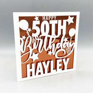 May include: White card with a copper-colored background. The card has a cut-out design that says "Happy 50th Birthday Hayley" with stars, balloons, and a party hat.