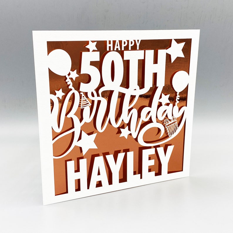 May include: White card with a copper-colored background. The card has a cut-out design that says "Happy 50th Birthday Hayley" with stars, balloons, and a party hat.