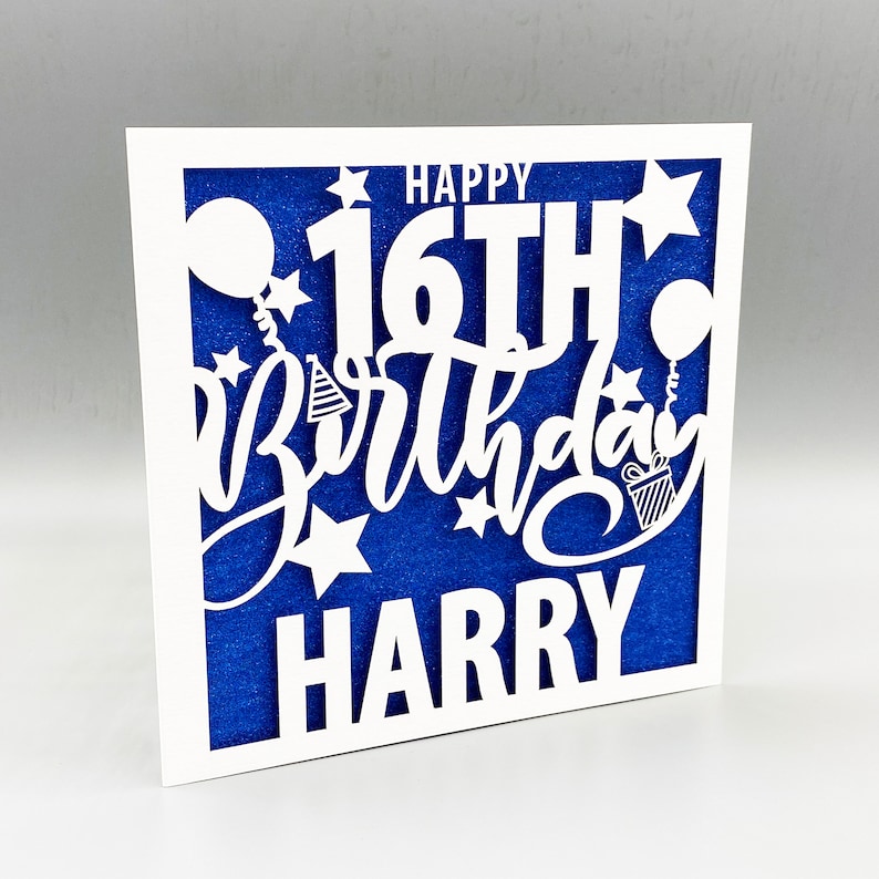 May include: White paper card with a blue background and a white cut-out design that says "Happy 16th Birthday Harry". The design includes stars, balloons, and a party hat.