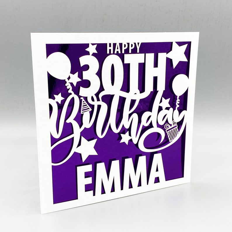 May include: A white card with a purple background and a white cutout design that says "Happy 30th Birthday Emma". The design includes stars, balloons, and a gift box.