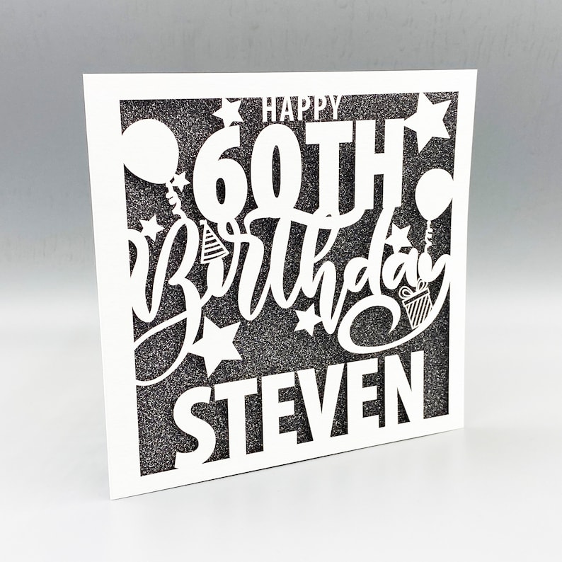 May include: A white card with a black glitter background and a white cut-out design that says "Happy 60th Birthday Steven".