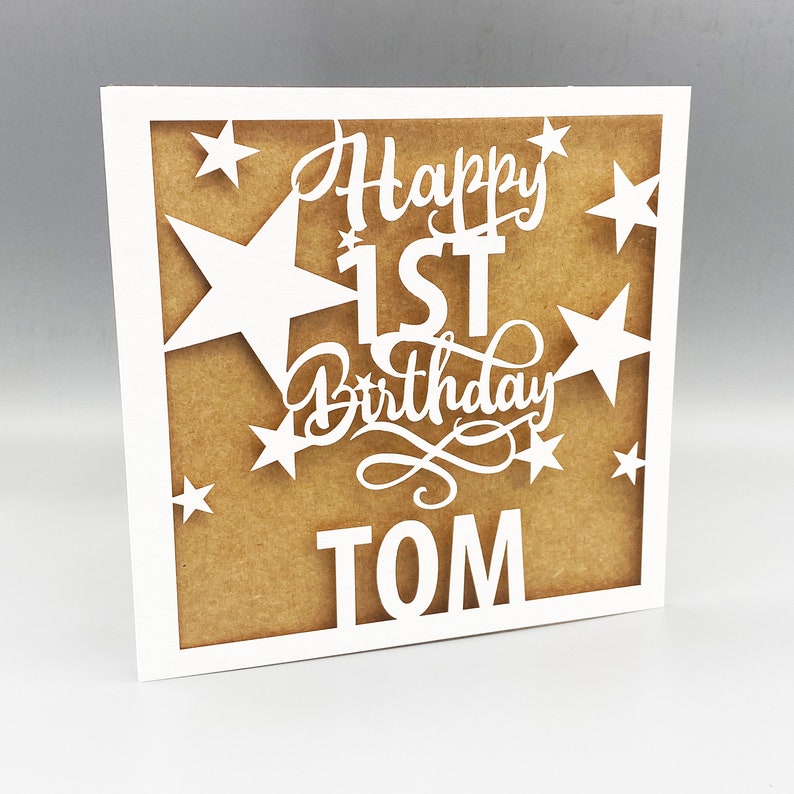 May include: White card with a brown background and a white cutout design. The design says "Happy 1st Birthday TOM" with stars around the text.
