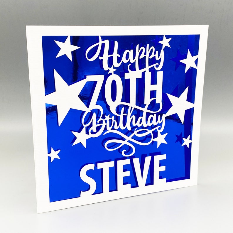 May include: White card with a blue background and white cut-out stars and the text "Happy 70th Birthday STEVE".