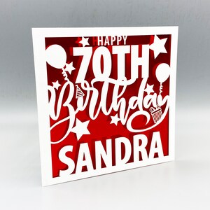 May include: White card with a red background and white cut-out lettering that says "Happy 70th Birthday Sandra" with stars and balloons.