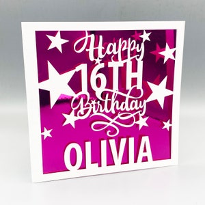 May include: A white card with a pink background and a white frame. The card has a white cutout design of stars and the text "Happy 16th Birthday Olivia".