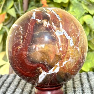 May include: A polished red, white, and brown agate sphere on a wooden stand. The sphere has a swirling pattern of colors and white veins.