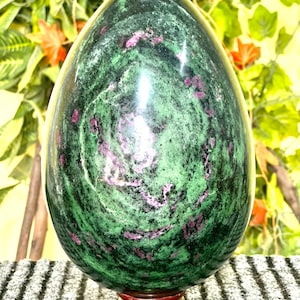 May include: A green and pink gemstone egg with a swirling pattern. The egg is sitting on a wooden stand.