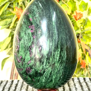 Puede incluir: Un huevo de piedra preciosa verde y rosa sobre un soporte de madera. El huevo está pulido y tiene una superficie lisa. La piedra preciosa es un tipo de zoisita.