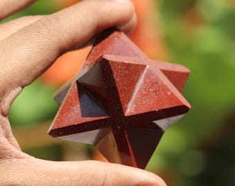 May include: A faceted, reddish-brown gemstone Merkaba star held in a hand. The star has multiple triangular faces that meet at points, creating a three-dimensional shape. The background is blurred with green and orange hues.