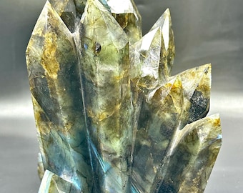 May include: A carved labradorite crystal cluster. The gemstone features a mix of green, blue, and gold hues. The crystal has multiple pointed tips and a textured surface, showcasing natural patterns.