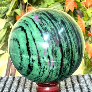 Pode incluir: Uma esfera de pedra preciosa verde, preta e rosa em um suporte de madeira. A esfera tem um padrão de redemoinho de preto e verde com acentos rosa.