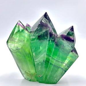 May include: A large, faceted fluorite crystal sculpture. The crystal features a vibrant green hue with streaks of purple and a polished, reflective surface. The geometric form has sharp, pointed edges and a layered appearance.