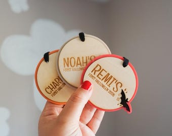 May include: Three circular wooden ornaments with coloured acrylic borders. Each ornament features engraved text including names like Noah, Remi, and Chari, along with the phrase First Halloween 2026. One ornament includes a small black bat and star icons.