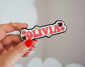 May include: A keychain with the name "OLIVIA" in red, retro-style lettering. The keychain has a black border and is decorated with flowers, ladybirds, and yellow stars. A gold-coloured keyring is attached.