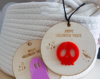May include: Two circular wooden tags with black string. One tag features a purple acrylic ghost and the text MYLA'S HALLOWEEN TREATS. The other tag features a red acrylic skull and the text JOEY'S HALLOWEEN TREATS, along with engraved star and bat icons.