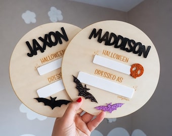 May include: Two circular light-coloured wooden plaques featuring the names JAXON and MADDISON in black 3D lettering. Each plaque includes the text HALLOWEEN and DRESSED AS with white rectangular spaces for writing, accented by glittery bat and skull shapes.