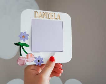 May include: A white picture frame with the name "DANIELA" in gold lettering. The frame is decorated with purple flowers and a cherry design. The frame is held by a hand with red nail polish.