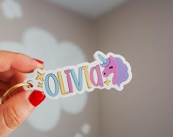 May include: A custom keyring featuring the name OLIVIA in colourful block letters with a purple unicorn head, a light blue horn, and small yellow stars and hearts. The design is printed on a white base and attached to a gold-toned metal ring.