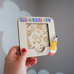 May include: A square wooden frame featuring the name MISS BRADLEY in pastel 3D letters at the top. The centre panel displays engraved school-themed icons like apples and pencils. A small pencil-shaped accent is attached to the right side.