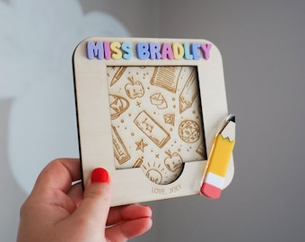 May include: A square wooden frame featuring the name MISS BRADLEY in pastel 3D letters at the top. The centre panel displays engraved school-themed icons like apples and pencils. A small pencil-shaped accent is attached to the right side.