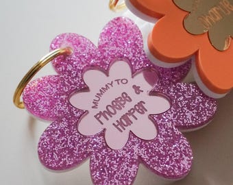 May include: Two flower-shaped keychains with gold-tone metal rings. One is pink with glitter and features the text 'MUMMY TO Phoebe & Harper'. The other is orange with a white base and a gold-tone centre, featuring the engraved name 'Charlie'.