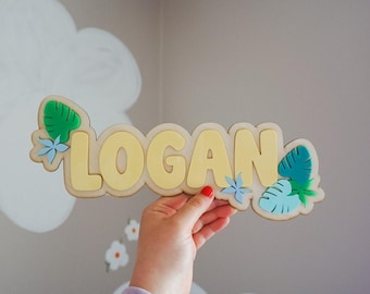 May include: A wooden name sign with the name "LOGAN" in yellow letters. The sign is decorated with green and blue leaf and flower accents. The sign is held up in front of a wall with cloud and flower decals.