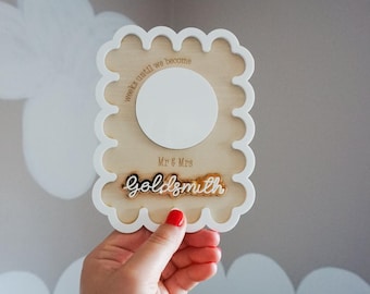 May include: A rectangular wooden sign with a scalloped white border featuring the engraved text weeks until we become Mr & Mrs and the name Goldsmith in raised white script lettering. A blank white circular space is centred on the wood surface.