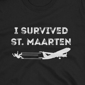 May include: Black t-shirt with white text that reads "I survived St. Maarten" and a silhouette of a plane flying low over a beach with a person running away from the plane.