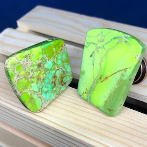 Pode incluir: Dois cabochões de pedra verde e branca marmorizada, um quadrado e outro retangular, sobre uma superfície de madeira clara.