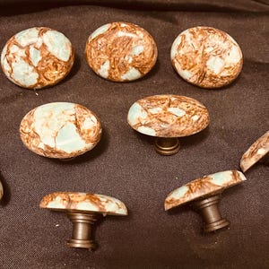 May include: A collection of eight decorative cabinet knobs. The knobs have a marbled appearance with brown and light blue tones. The hardware is a dark bronze color. The knobs are round and oval shaped.