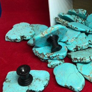 May include: Turquoise stone cabinet knobs with black fittings. The knobs are a vibrant turquoise colour with brown veining, and are scattered on a red surface. Some knobs are spilling out of a cardboard box.