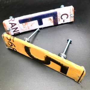 May include: Two vintage license plates repurposed as drawer pulls. The top plate is white with blue and black lettering, reading "C-PANI" and "F". The bottom plate is yellow with black lettering, reading "566" and "5". Both plates have screws and metal hardware.