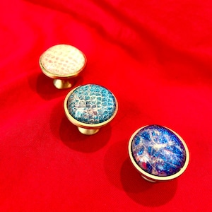 May include: Three decorative cabinet knobs with brass bases and colorful, patterned tops. One knob has a white, textured surface, while the others feature blue and iridescent designs. The knobs are set against a vibrant red background.