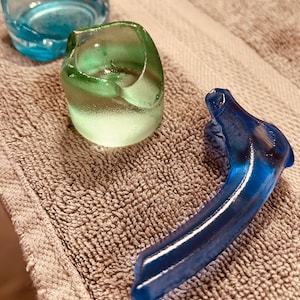 May include: Three pieces of colorful glass art. A blue ashtray, a green ring-shaped object, and a blue curved handle. The objects are on a brown towel.