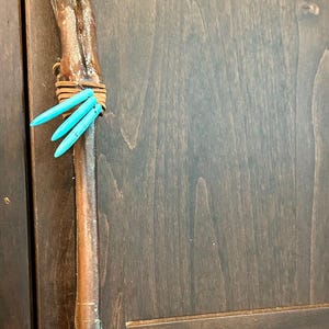 May include: A handcrafted walking stick made from a deer leg bone, adorned with turquoise-colored accents. The stick is wrapped with leather strips and has a dark brown finish. The background is a dark wood cabinet.