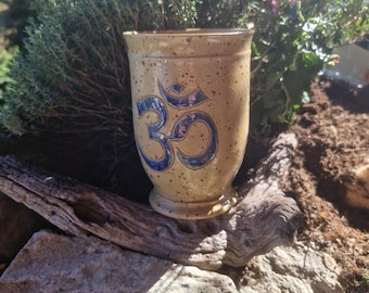 Könnte beinhalten: Eine beige Keramiktasse mit einem blauen Om-Symbol. Die Tasse hat eine gesprenkelte Oberfläche und einen ausgestellten Rand. Sie steht auf einer natürlichen Holz- und Steinoberfläche, mit grünem Laub im Hintergrund.