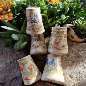 Könnte beinhalten: Fünf kleine Keramikbecher mit Blumen- und Traubenmotiven. Die Becher sind weiß mit handbemalten Details in Blautönen, Grün, Rot und Gelb.