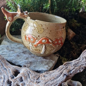 Op de afbeelding: Een gespikkelde beige keramische mok met een reliëfontwerp van oranje en witte paddenstoelen aan de buitenkant. Het handvat is gevormd als een klein dier met puntige oren, wat een sculpturaal element toevoegt aan het drinkgerei.