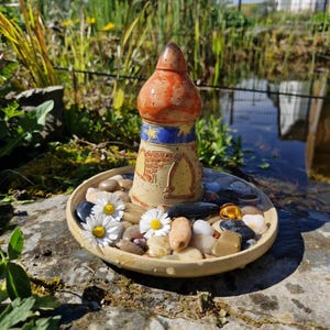 Peut inclure: Ornement de jardin en céramique en forme de phare, avec un dessus marron et orange, des accents bleus et jaunes et un motif en forme de brique. Il repose sur une coupelle peu profonde remplie de galets et de marguerites.