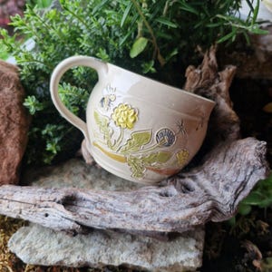 Può includere: Tazza in ceramica color crema con manico curvo, decorata con fiori gialli in rilievo, foglie verdi e un'ape. La tazza è appoggiata su uno sfondo di pietra naturale e legno, con vegetazione.