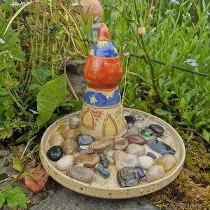 Könnte beinhalten: Ein skurriles Vogelbad aus Keramik mit einem farbenfrohen, turmartigen Design. Der Turm ist orange und blau mit Sternakzenten. Die Basis ist eine flache Schale, gefüllt mit Kieselsteinen, ideal für einen Garten oder Außenbereich.