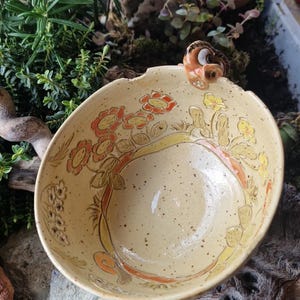 Könnte beinhalten: Eine handgefertigte Keramikschale mit hellbeiger Glasur. Die Schale hat ein Blumenmuster in Orange, Gelb und Braun, mit einer skurrilen vogelartigen Figur am Rand. Ideal zum Servieren von Speisen oder als dekoratives Stück.
