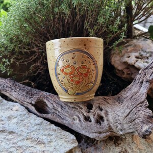 Könnte beinhalten: Ein beigefarbener Keramik-Blumentopf mit floralem Design. Der Topf hat ein kreisförmiges Design mit roten und orangen Blumen, grünen Blättern und einem blauen Rand. Der Topf steht vor einem natürlichen Hintergrund aus Holz, Felsen und Grün.