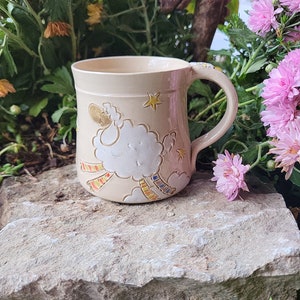 Puede incluir: Una taza de cerámica beige con un diseño caprichoso de una oveja volando a través de las nubes con rayas de colores. La taza tiene un asa y está decorada con estrellas.