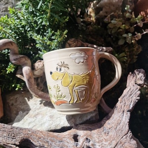 Op de afbeelding: Crèmekleurige keramische mok met een cartoon hond ontwerp in geel en groen. De mok heeft een gebogen handvat en een wolkenillustratie. De mok staat tegen een natuurlijke achtergrond met groen en hout.
