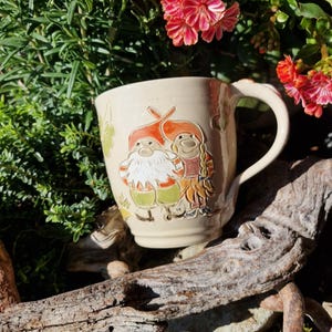 Könnte beinhalten: Cremefarbene Keramiktasse mit einem skurrilen Zwergen-Design. Die Tasse hat einen gebogenen Henkel und ist mit zwei Zwergen mit roten Hüten und grünen Hosen verziert. Der Hintergrund umfasst Grün, Blumen und ein Stück Treibholz.