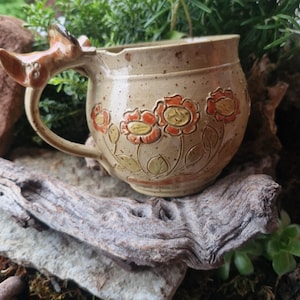 Könnte beinhalten: Beige Keramikbecher mit hirschförmigem Griff und geprägtem Blumendesign. Die Blumen sind orange mit gelben Zentren und grünen Blättern. Der Becher steht auf einem Stück Treibholz.