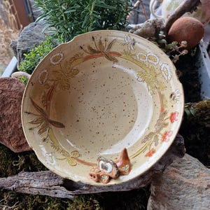 Op de afbeelding: Een beige keramische kom met een bloemmotief in geel, oranje en bruin. De kom heeft een getextureerd oppervlak en een decoratief uilfiguur aan de rand. De kom staat op een bedje van mos en hout.