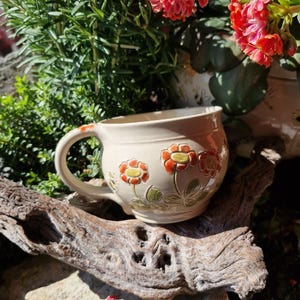 Könnte beinhalten: Eine beige Keramiktasse mit großem Henkel, verziert mit orangefarbenen und gelben Blumenmustern. Die Tasse steht auf einem Stück Treibholz, mit Grün und roten Blumen im Hintergrund.